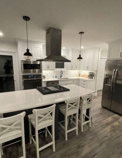 newly renovated kitchen with gleaming white cabinetry and countertops, a large island with navy cabinetry, and a central stainless steel range hood.